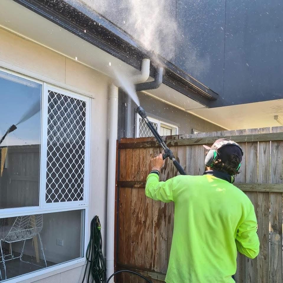 Michael in Brisbane wearing a high-visibility shirt pressure washing a house exterior and eaves near a wooden fence.