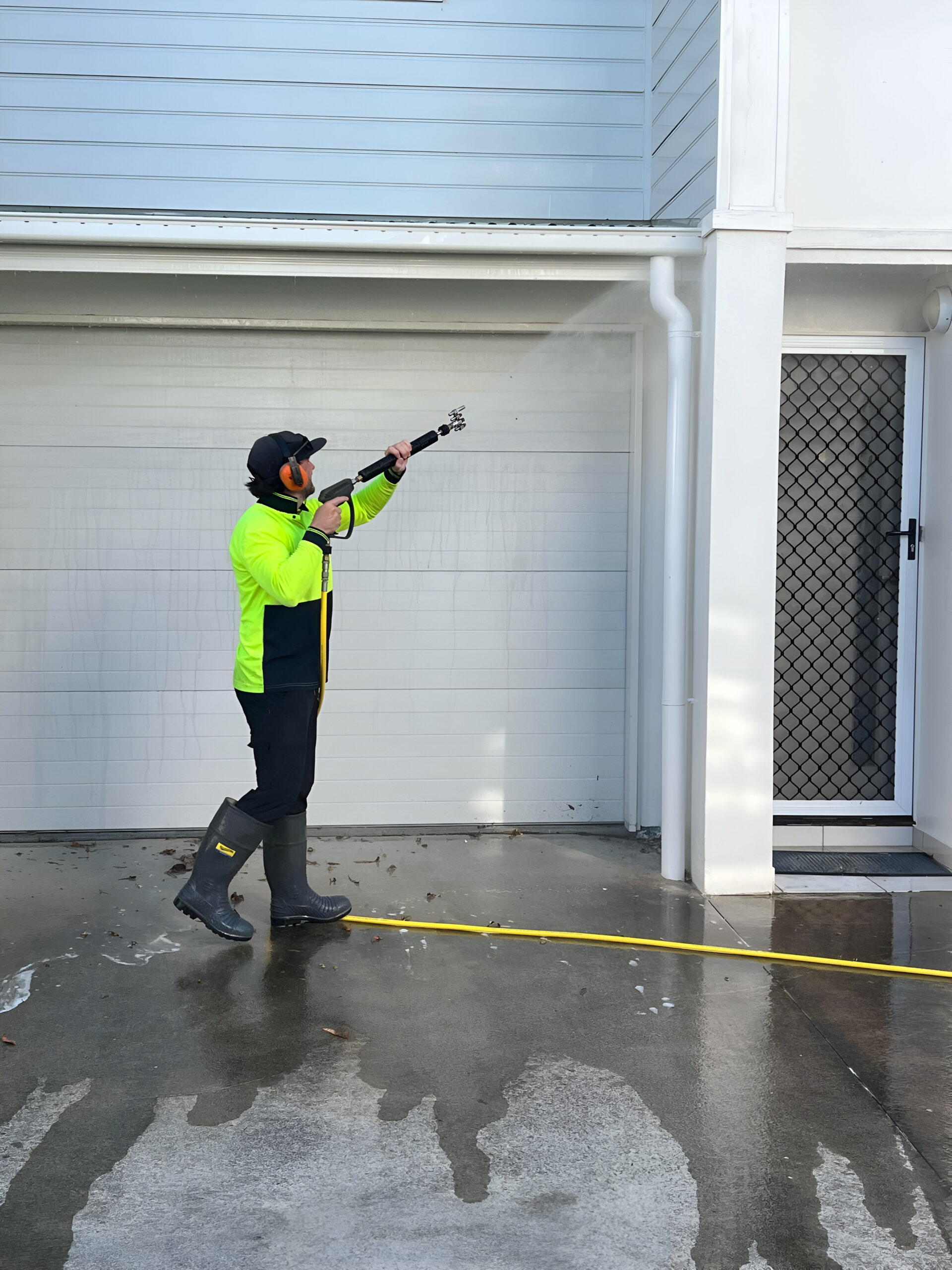 Michael working in Caboolture wears safety gear while using a pressure washer to clean the exterior wall near the garage door and entrance of a house.