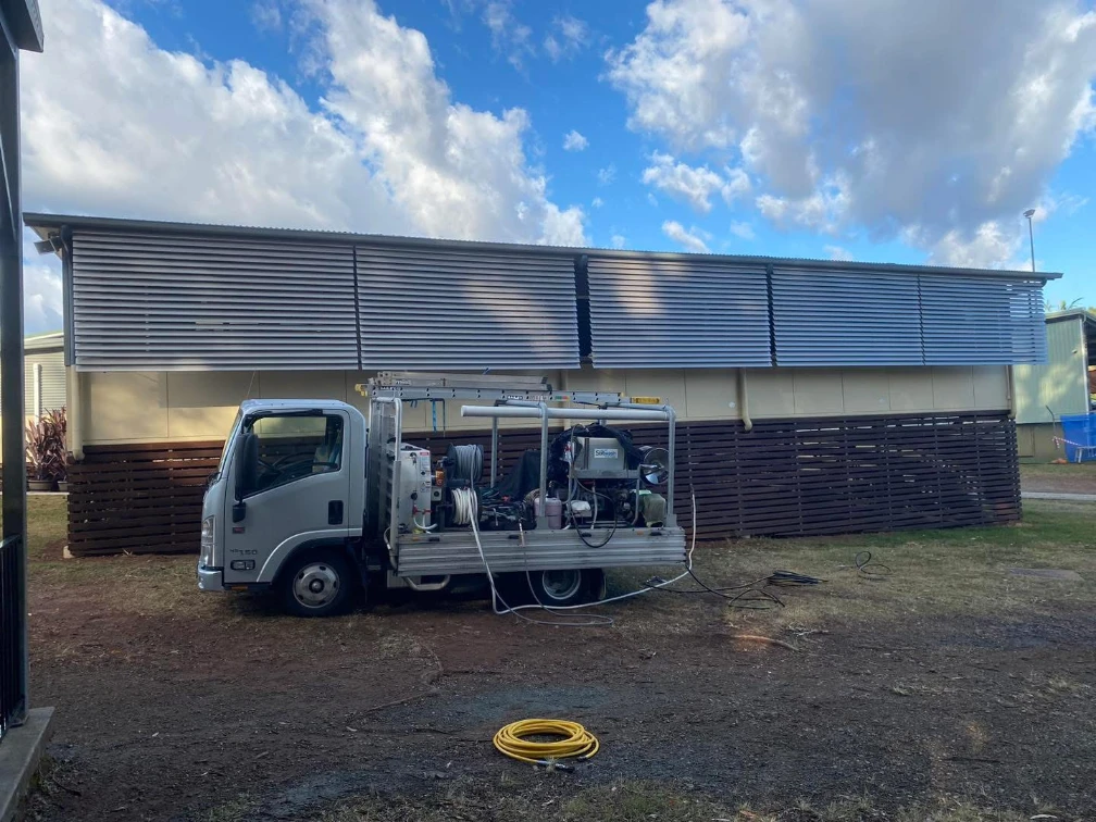 QQCPM utility truck with cleaning equipment parked beside a building with metal siding and wood panels on a cloudy day.